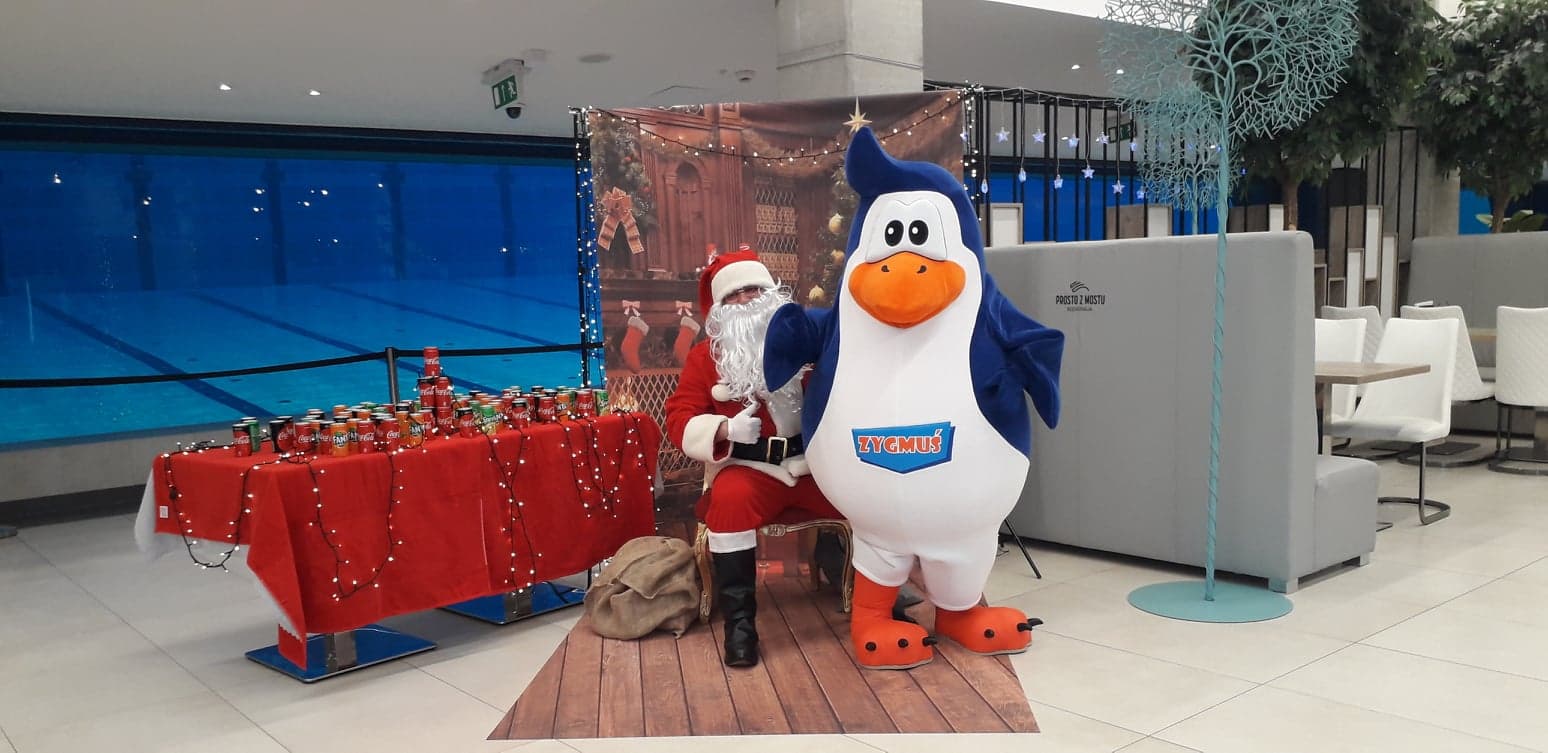 Do zdjęcia pozuje strój reklamowy w postaci Pingwina Zygmusia w towarzystwie Świętego Mikoła. Fotografia wykonana zapewne w foyer centrum handlowego.